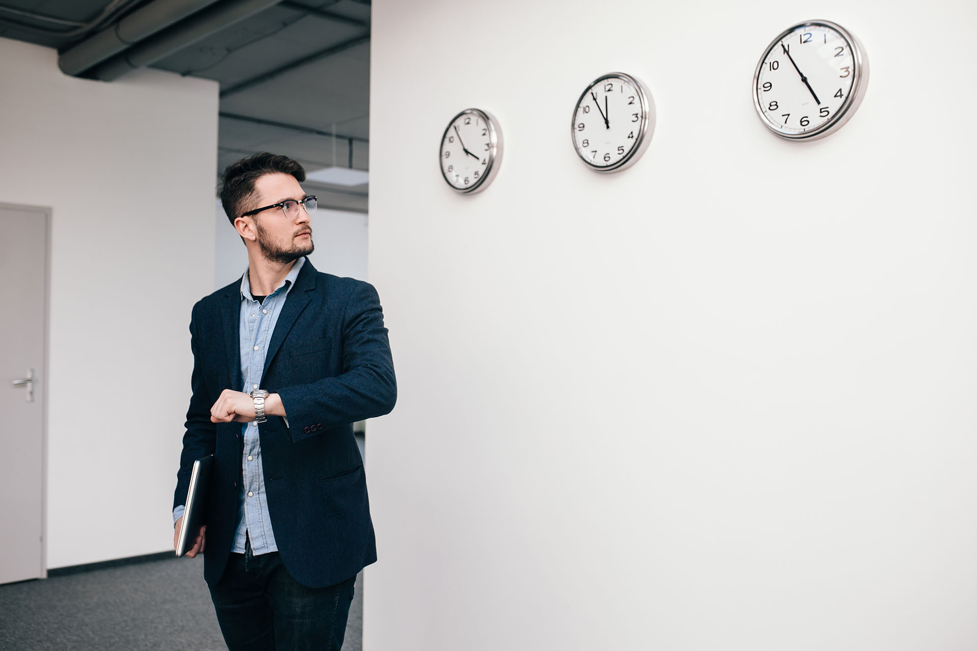 Young guy in glasses is  walking in office. He wears blue shirt, dark jacket, jeans and beard. He holds  laptop in hand. He is looking to clock on wall