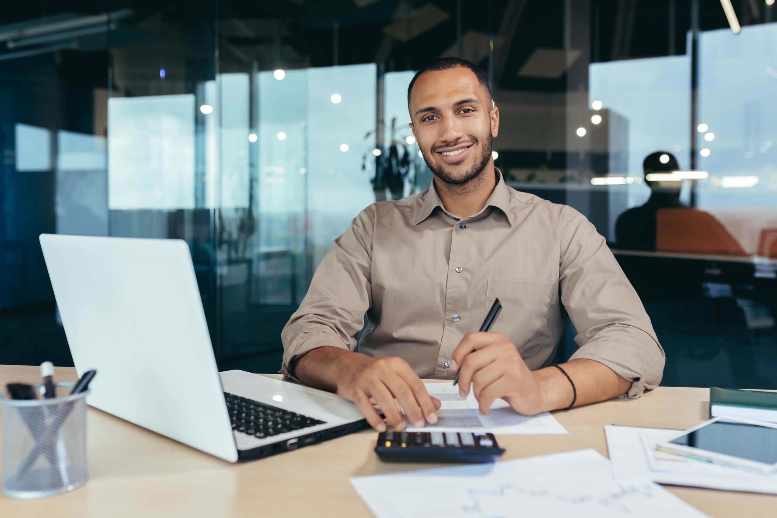 Portrait of young successful hispanic businessman inside office, man smiling and looking at camera, paper worker happy with achievement results sitting at workplace with laptop